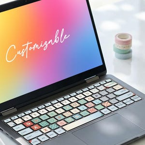 Puede incluir: Un portátil gris oscuro con una pantalla degradada que muestra la palabra Customizable en letra cursiva blanca. El teclado tiene teclas cubiertas con pegatinas estampadas de varios colores, letras y diseños. Una pequeña figura de perro está cerca.
