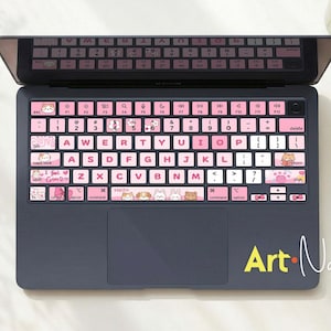 May include: A laptop with a pink and white keyboard cover featuring cartoon animal designs. The keyboard cover is on a dark gray laptop. The image also includes a vase of flowers and a small cat figurine.