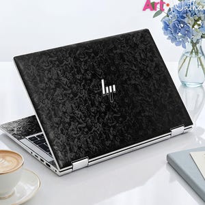Op de afbeelding: Een laptop met een zwarte, gemarmerde skin op de deksel en een zilverkleurig HP-logo in het midden. De laptop staat open en toont een zwart toetsenbord en een zilverkleurig frame. Het apparaat staat op een witte ondergrond naast een kop koffie.