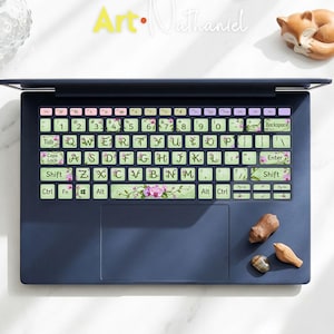 Könnte beinhalten: Ein marineblauer Laptop mit floralen Tastaturaufklebern. Die Tastaturtasten sind grün mit rosa Blumen und Blättern. Der Text "Art Nathaniel" ist in Gelb und Weiß gehalten. Eine kleine Fuchsfigur und andere dekorative Gegenstände befinden sich in der Nähe.