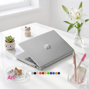 May include: A silver laptop with a textured carbon fiber pattern on the lid and a centered HP logo. The laptop is surrounded by a small succulent in a white planter, a cat figurine, a clear pen holder with pens, and a white lily in a glass vase.