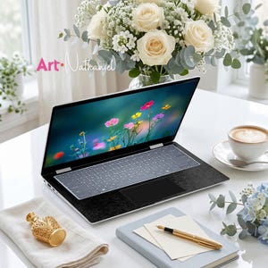 Op de afbeelding: Een zilveren laptop met een zwarte, getextureerde polssteun en een scherm met kleurrijke wilde bloemen. Daarnaast staan een gouden parfumflesje op een witte doek, een lichtblauw notitieboek met een gouden vulpen en een kopje koffie op een schoteltje.