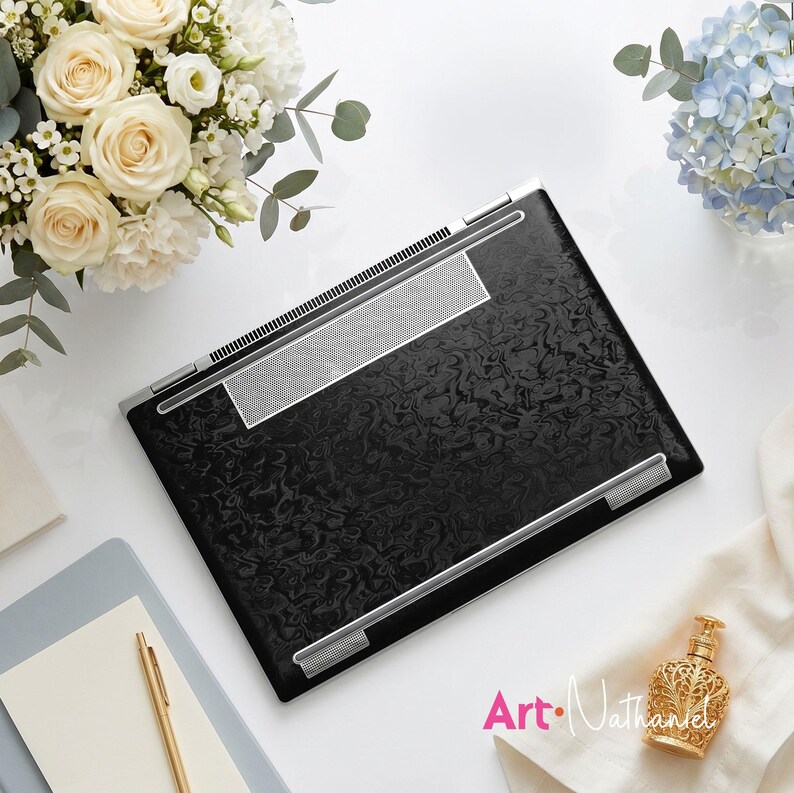 Op de afbeelding: Een gesloten laptop met een zwarte, getextureerde skin met een wervelend, marmerachtig patroon. Het apparaat heeft zilverkleurige metalen scharnieraccenten en een rechthoekig zilverkleurig luidsprekerrooster langs de bovenrand.