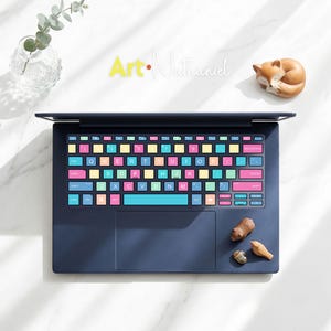 May include: A dark blue laptop featuring a custom keyboard skin with multi-colored keys in shades of pink, yellow, light blue, and green. The keys display white letters, numbers, and symbols. The laptop has a smooth trackpad and a dark blue frame.