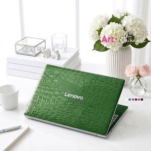 May include: A Lenovo laptop with a textured green crocodile skin pattern on the lid. The laptop is closed and positioned on a white surface. The design features a realistic reptilian scale texture in a deep emerald green color with the white Lenovo logo.