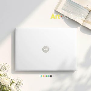 May include: A white Dell laptop with a closed lid featuring the centered silver Dell logo. The laptop is positioned near an open book with a coastal illustration and a small cluster of white flowers. A row of small colored circles is visible.