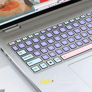 K&ouml;nnte beinhalten: Ein silberner Laptop mit einer individuellen Tastaturabdeckung im Color-Blocking-Design. Die Tasten sind in Hellblau, sanftem Violett und Blassrosa gehalten und mit schwarzer Schrift versehen. Das Layout umfasst Standard-QWERTY-Tasten.