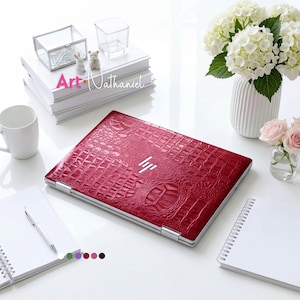 May include: A closed laptop featuring a deep red, textured crocodile skin pattern on its outer lid with a silver HP logo in the centre. The device is positioned on a white surface surrounded by notebooks, a pen, a white mug, and a vase of flowers.