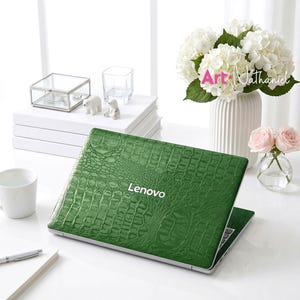 Puede incluir: Un ordenador portátil Lenovo con una pegatina de textura de piel de cocodrilo verde vibrante en la tapa exterior. La superficie muestra un patrón detallado de escamas de reptil en relieve en toda la cubierta superior, con el logo Lenovo blanco.