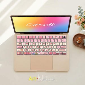 Könnte beinhalten: Ein hellbeiges Laptop mit einer rosa Tastaturabdeckung mit weißen Tasten mit rosa Schrift und Cartoon-Illustrationen. Der Bildschirm zeigt das Wort "Customizable" in weißer Schrift vor einem farbenfrohen Hintergrund. Der untere Teil des Bildes trägt den Text "Art-Nathaniel".