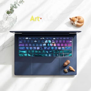 May include: A dark blue laptop featuring a keyboard skin with a printed landscape illustration of mountains, clouds, and a sunset. The keys display white letters and symbols. The laptop is positioned on a white surface with decorative items.