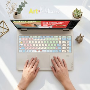 May include: A beige laptop with a colorful keyboard cover featuring pastel-colored keys, small cartoon illustrations of cats, mushrooms, and watermelons, and the text Art-Nathaniel displayed above the screen on a white surface.