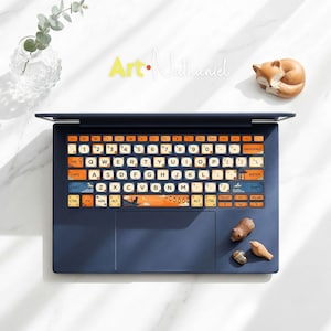 May include: A navy blue laptop with decorative keyboard stickers featuring a landscape design. The keyboard keys are white with orange accents. The image also includes a small fox figurine and the text "Art Nathaniel".