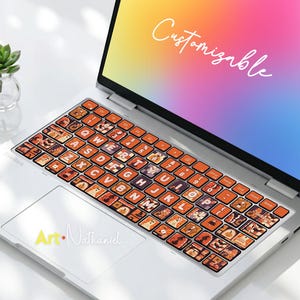 May include: A silver laptop featuring a keyboard skin with an orange Halloween-themed pattern of ghosts, pumpkins, and bats. The screen displays the word Customizable in white script, and the trackpad features the text Art Nathaniel in yellow and white.