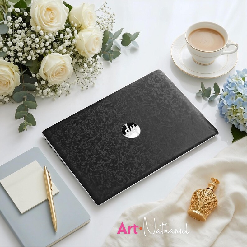 Op de afbeelding: Een zwarte laptop met een gestructureerd, wervelend patroon op het deksel en een cirkelvormig zilveren logo in het midden. De laptop is omringd door witte rozen, gipskruid, een kopje koffie, een lichtblauw notitieboek, een gouden pen en een gouden flesje.