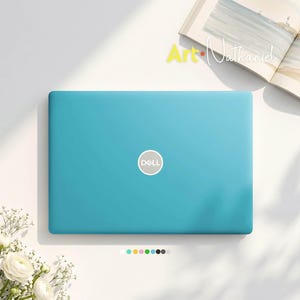 May include: A bright turquoise Dell laptop with a centered silver logo on the lid. The laptop is positioned near an open book with the text Art Nathaniel visible on the page. A small row of colored circular icons is located below the laptop.