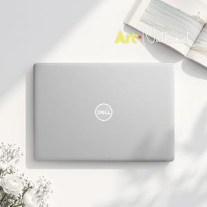 May include: A silver Dell laptop with a closed lid featuring the white Dell logo in the centre. The laptop is positioned near an open book with the text Art Nathaniel visible on the page and a cluster of white flowers in the corner.