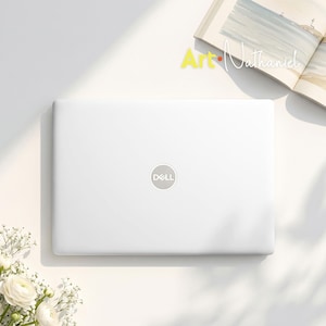 Puede incluir: Un ordenador portátil Dell blanco con la tapa cerrada que muestra el logotipo plateado de Dell en el centro. El portátil está situado junto a un libro abierto con una ilustración costera y un pequeño ramo de flores blancas con tallos verdes.