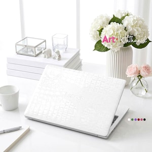 May include: A white Lenovo laptop with a textured crocodile skin pattern on the lid. The laptop is closed and positioned on a white surface. The brand name Lenovo is embossed in the centre of the lid in a matching white finish.