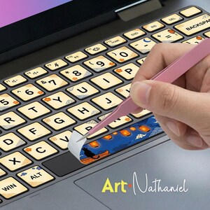 Puede incluir: Teclado de ordenador portátil con pegatinas decorativas que se están aplicando. Las pegatinas presentan un diseño azul con linternas naranjas y una silueta negra. Las teclas del teclado son beige con letras negras y detalles florales naranjas.