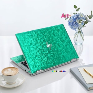 May include: A laptop with a vibrant emerald green marbled skin featuring a metallic sheen and a centered silver HP logo. The device is open, displaying a textured surface pattern that mimics swirling liquid, set against a clean white background.