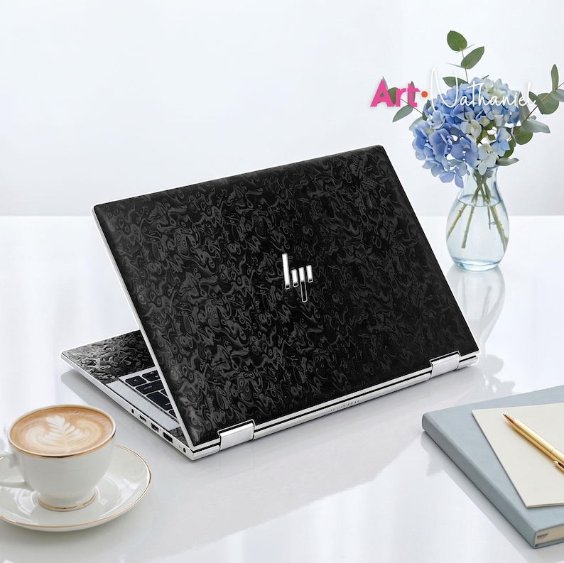 Op de afbeelding: Een laptop met een zwarte, gemarmerde skin op de deksel en een zilverkleurig HP-logo in het midden. De laptop staat open en toont een zwart toetsenbord en een zilverkleurig frame. Het apparaat staat op een witte ondergrond naast een kop koffie.