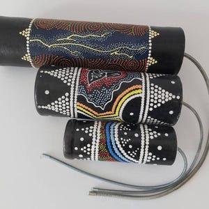 Op de afbeelding: Drie zwarte didgeridoos met kleurrijke stippenkunstontwerpen. De grootste heeft een blauwe achtergrond met gele en rode stippen. De middelste heeft een rood en wit ontwerp. De kleinste heeft een blauw en rood ontwerp. Alle zijn verbonden door een metalen veer.