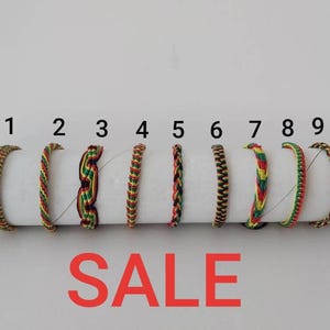 May include: Nine colourful woven friendship bracelets in various patterns and colours, including red, yellow, green, and black. The word "SALE" is displayed in large red letters below the bracelets.