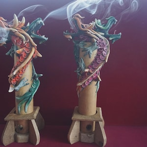 Può includere: Due bruciatori di incenso cilindrici con figure di drago scolpite avvolte attorno ai corpi verticali. I draghi sono dipinti in tonalità di rosso, arancione e verde. Ogni bruciatore poggia su una base in legno chiaro ed emette sottili fili di fumo.