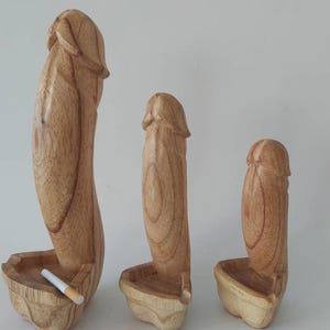 Peut inclure: Trois sculptures en bois en forme phallique de hauteurs variées, chacune dotée d'une base sculptée servant de cendrier. Le grain du bois brun clair naturel est visible sur toutes les surfaces lisses. Une cigarette blanche repose dans la plus grande.