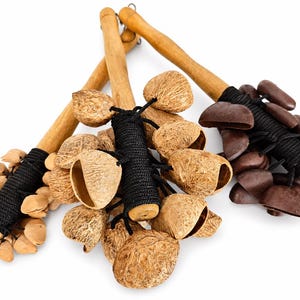 Puede incluir: Tres instrumentos de percusión con mangos de madera y varias vainas de semillas adjuntas. Las vainas de semillas son en tonos marrones y beige, y están unidas a los mangos con cordón negro.