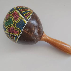 May include: A maraca featuring a rounded, dark brown coconut shell body with a smooth, light-colored wooden handle. The upper portion of the shell is decorated with a vibrant, hand-painted geometric pattern of dots in yellow, red, green, and blue.