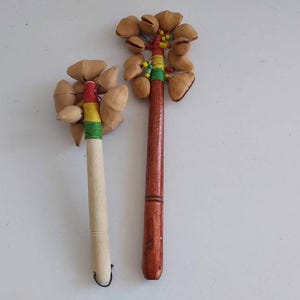 Könnte beinhalten: Zwei handgefertigte Percussion-Instrumente. Eines hat einen hellen Holzgriff mit roten, gelben und grünen Akzenten, das andere einen dunklen Holzgriff. Beide haben Samenkapseln oben befestigt. Die Instrumente befinden sich auf einer weißen Oberfläche.