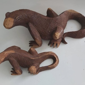 May include: Two intricately carved wooden lizard sculptures. The lizards are brown with a textured, scale-like pattern. One lizard is larger than the other. The sculptures have detailed claws and curved tails, showcasing a naturalistic design.