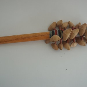 Può includere: Uno strumento a percussione in legno, probabilmente un sonaglio, con un lungo manico marrone chiaro. La testa presenta numerosi oggetti beige a forma di seme, attaccati con perline rosse e una fascia di tessuto fantasia.