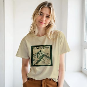 May include: A cream-coloured t-shirt with a mountain graphic inside a dark green square, and the text "ELEVATION 2026". The mountain illustration is in green and tan shades. The shirt has short sleeves and is made of a soft material.