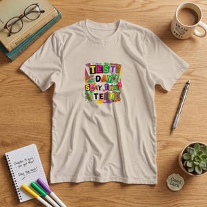 Puede incluir: Una camiseta color crema con el texto "TEST DAY SLAY THAT TEST" en letras coloridas y estampadas. La camiseta se muestra sobre una superficie de madera con libros, una taza y bolígrafos.