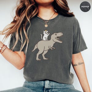 Puede incluir: Camiseta gris oscuro con una ilustración de dibujos animados de un gato blanco con corona montado en un Tyrannosaurus Rex marrón claro. La camiseta tiene un corte holgado y mangas cortas. La palabra "Pepper" está en un círculo en la esquina superior derecha.