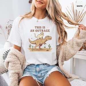 Puede incluir: Camiseta blanca con una ilustración de dibujos animados de un gato con corona montado en un dinosaurio, con el texto "THIS IS AN OUTRAGE" en marrón. El diseño incluye cactus del desierto. Una prenda casual.