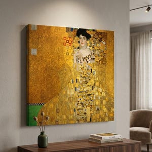 May include: A large, square canvas print of a Gustav Klimt painting. The artwork features a woman in a long, ornate gold dress with intricate patterns. The background is a textured gold, with touches of green and geometric shapes.