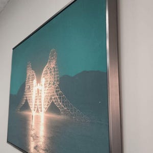 May include: A rectangular wall art piece featuring a teal-toned landscape with two large, glowing, wire-frame human figures facing each other. The artwork is encased in a thin, metallic silver frame and is mounted on a plain, light-colored wall.