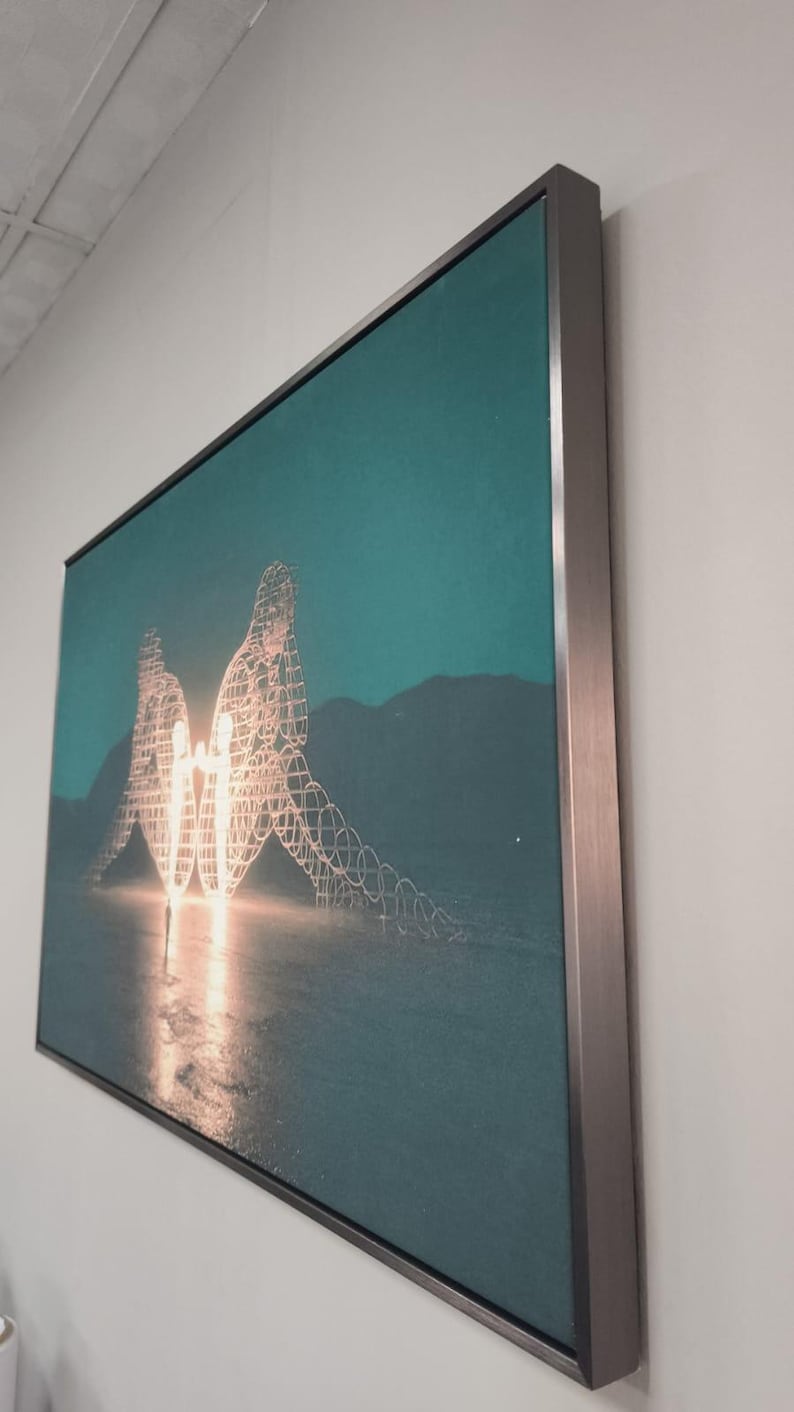 May include: A rectangular wall art piece featuring a teal-toned landscape with two large, glowing, wire-frame human figures facing each other. The artwork is encased in a thin, metallic silver frame and is mounted on a plain, light-colored wall.
