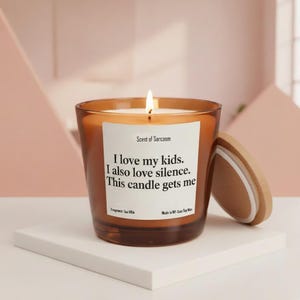 Peut inclure: Une bougie en verre brun allumée avec un couvercle en bois, posée sur une surface blanche. La bougie porte une étiquette blanche avec le texte "Scent of Sarcasm" et la citation "I love my kids. I also love silence. This candle gets me."