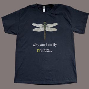 May include: A black short-sleeved t-shirt featuring a detailed illustration of a dragonfly with translucent wings. Below the insect, the text reads "why am i so fly" followed by the National Geographic logo in yellow and white lettering.