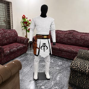 Puede incluir: Un disfraz blanco con un casco negro, con una parte superior acolchada, pantalones blancos y un cintur&oacute;n con munici&oacute;n. El disfraz incluye una funda marr&oacute;n y un delantal blanco con un emblema negro. El maniqu&iacute; est&aacute; sobre una base negra.