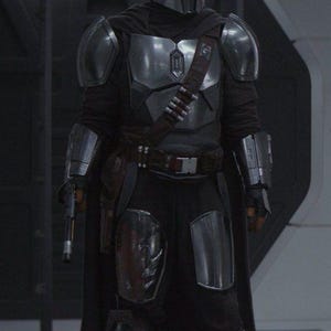 May include: A full-body shot of a character in a silver and black Mandalorian armor suit. The helmet is silver, and the armor covers the chest, shoulders, and forearms. A dark cape and belt with ammunition pouches complete the look.