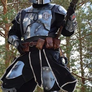 May include: A detailed costume of a Mandalorian warrior, featuring a helmet, chest armor, and utility belt. The armor is primarily blue and silver, with black accents. The warrior is holding a blaster rifle, set against a backdrop of trees.
