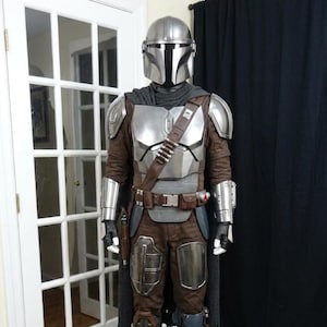May include: A full-body costume resembling a character from a science fiction series. The costume features a silver helmet, chest armor, and brown and silver accents. It includes a cape, belt, and boots, with a holster for a weapon.