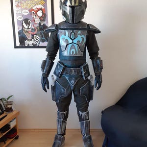 Puede incluir: Un disfraz de cuerpo completo que se asemeja a un guerrero mandaloriano. La armadura es principalmente gris oscuro con detalles en azul en la placa del pecho. El casco tiene una visera en forma de T y dos antenas. El disfraz incluye un traje interior negro, guantes y botas.