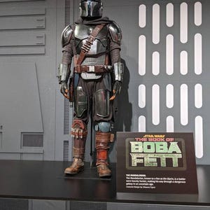 Puede incluir: Un disfraz completo de mandaloriano con casco, placa pectoral y hombreras de metal plateado. El traje incluye correas de cuero marrón oscuro, un cinturón utilitario y botas marrones. Un letrero dice Star Wars The Book of Boba Fett.